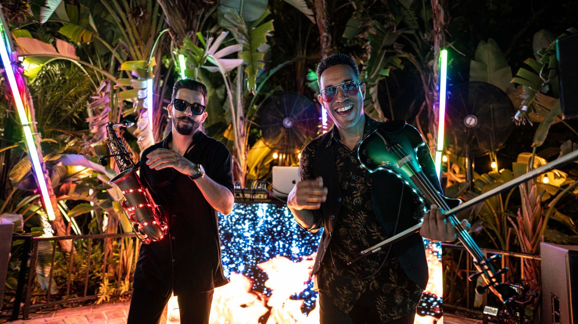 Two people posing at night in a colorful neon-lit garden, holding glowing props.
