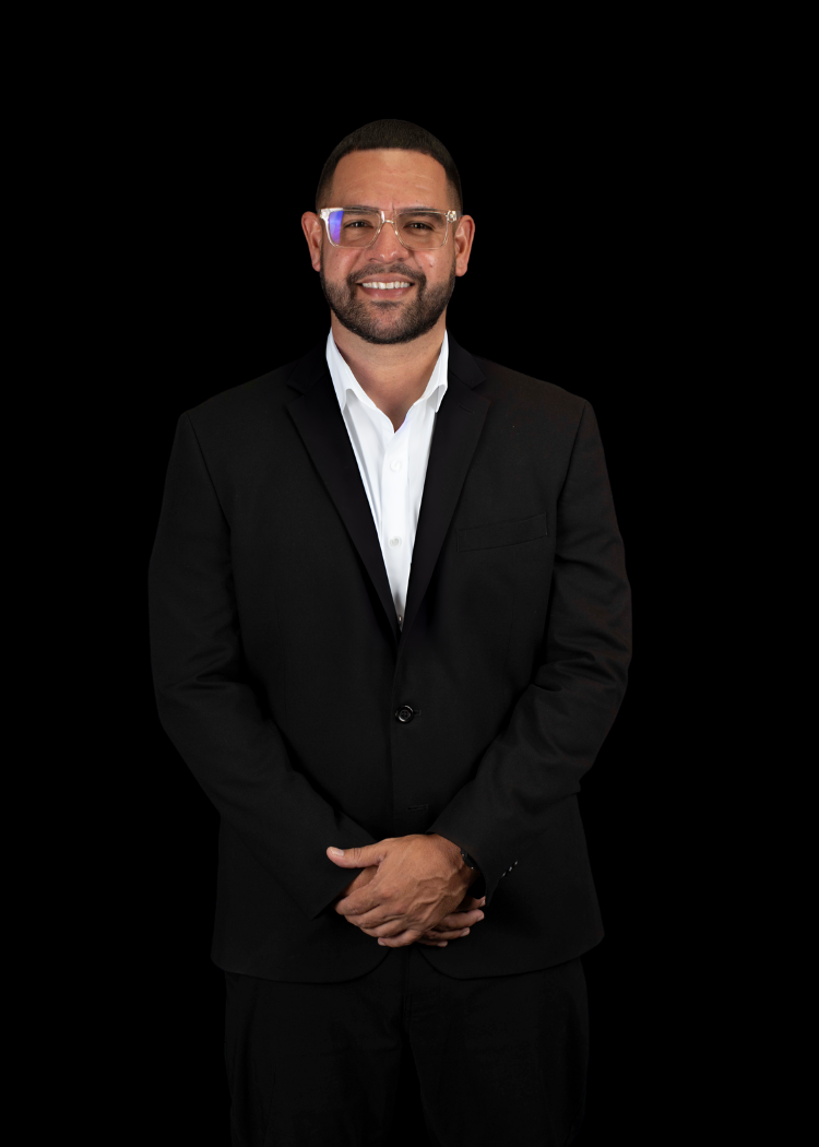 Man in a black suit and white shirt standing against a black background, hands clasped and smiling.