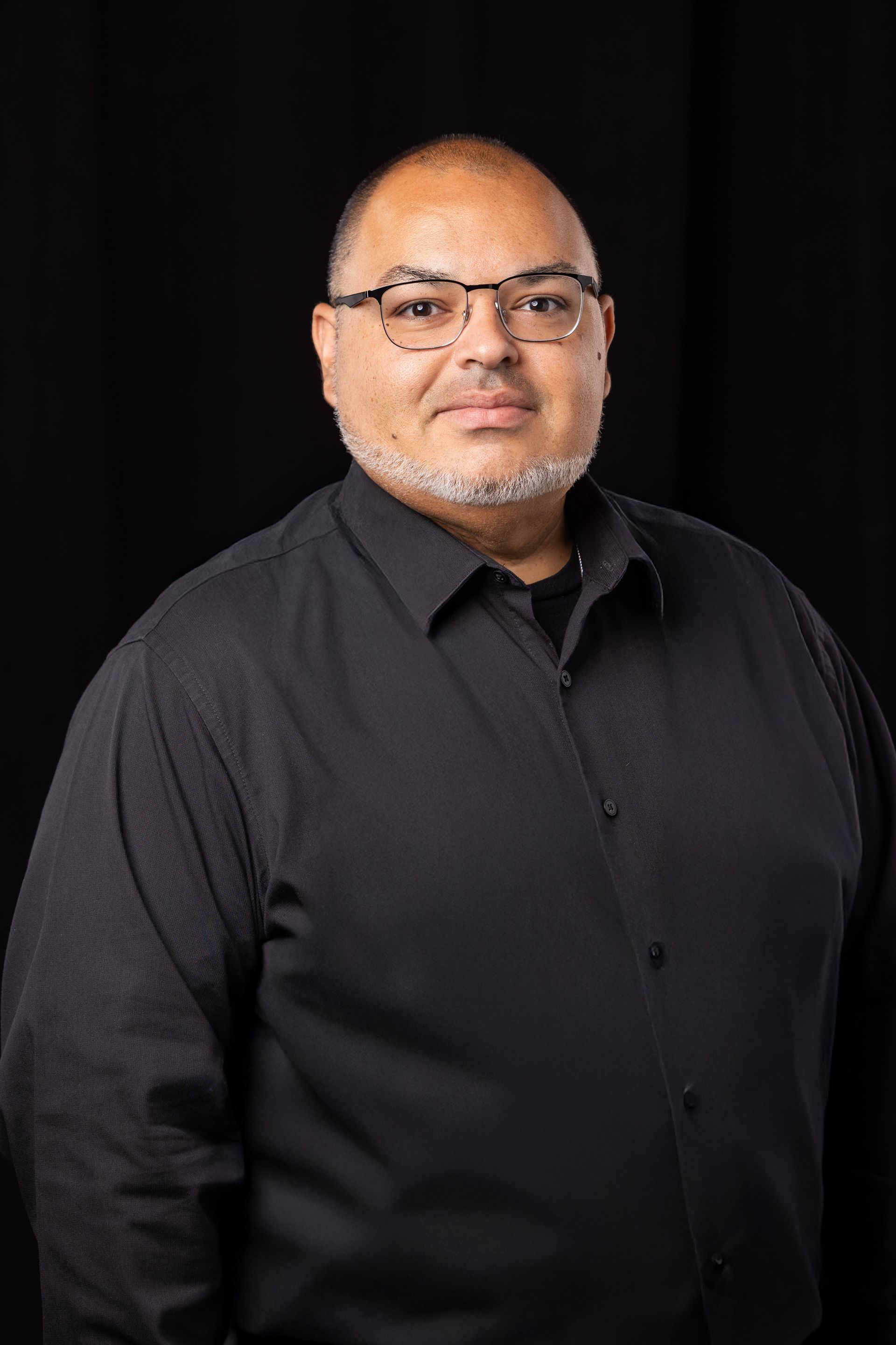 Portrait of a man in a black shirt against a dark background, facing the camera with a slight smile