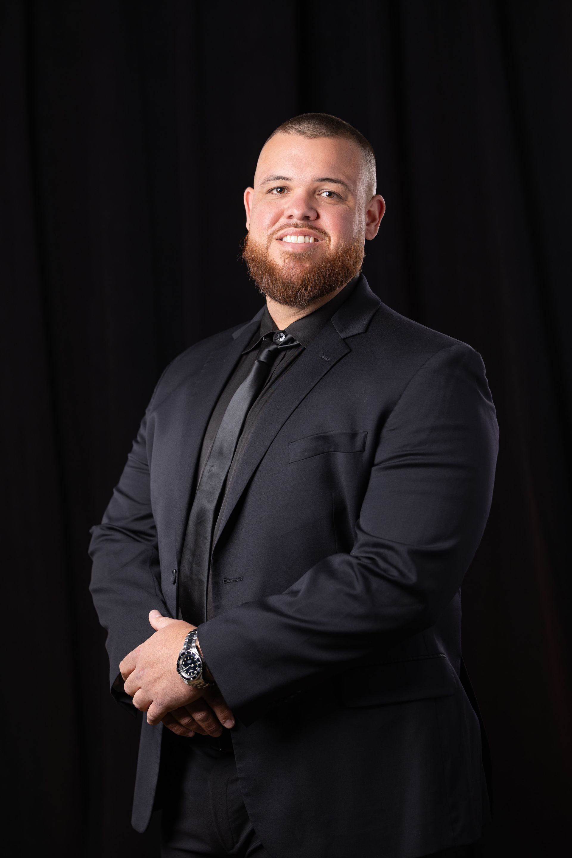 Man in a black suit posing against a dark backdrop, hands clasped and smiling