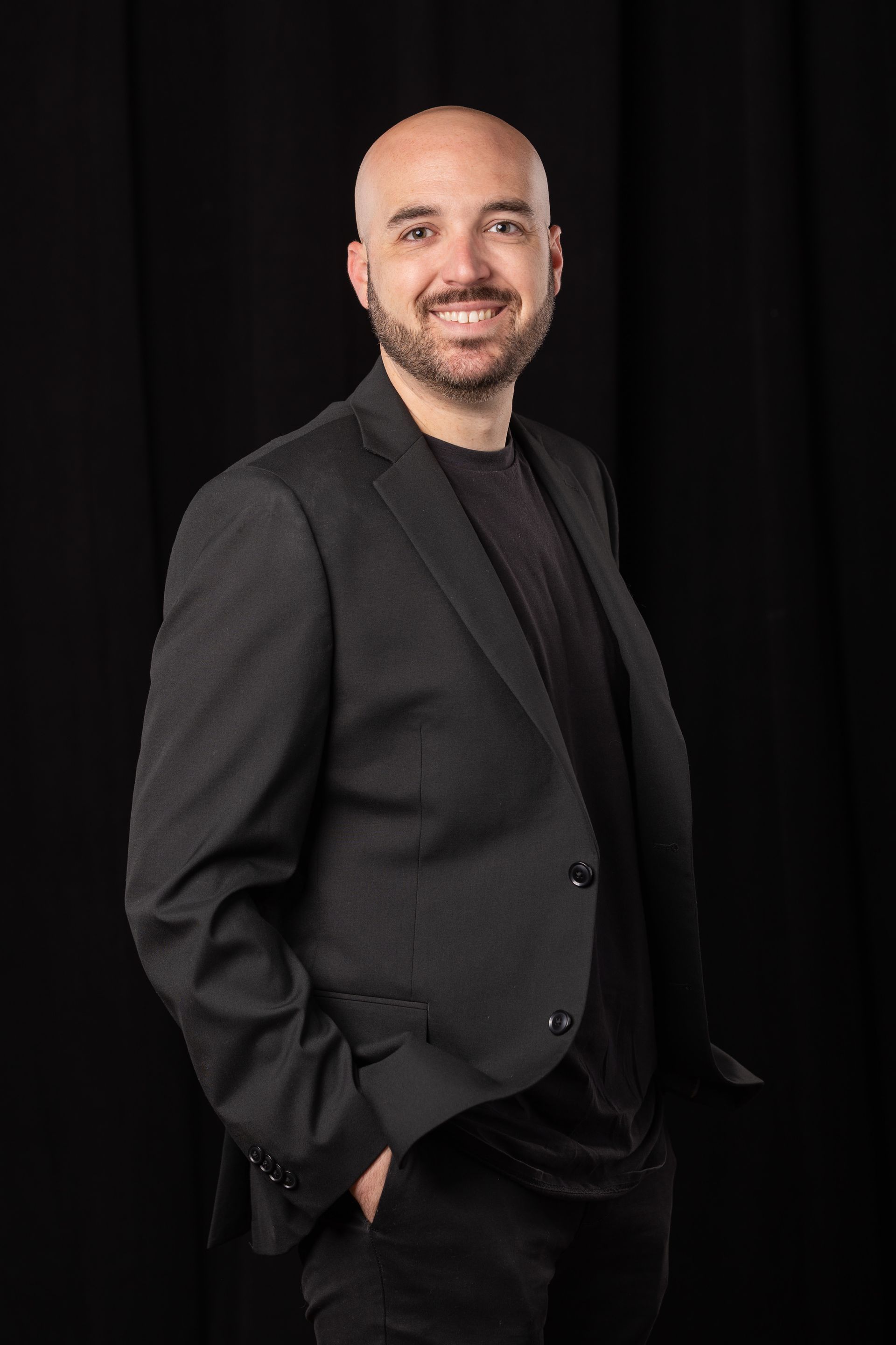 Man in a black suit smiling against a dark studio background, hands in pockets.