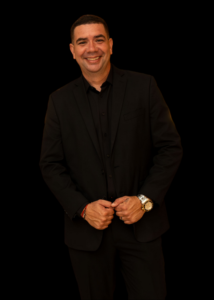 Smiling man in black suit standing against a black background, holding his jacket