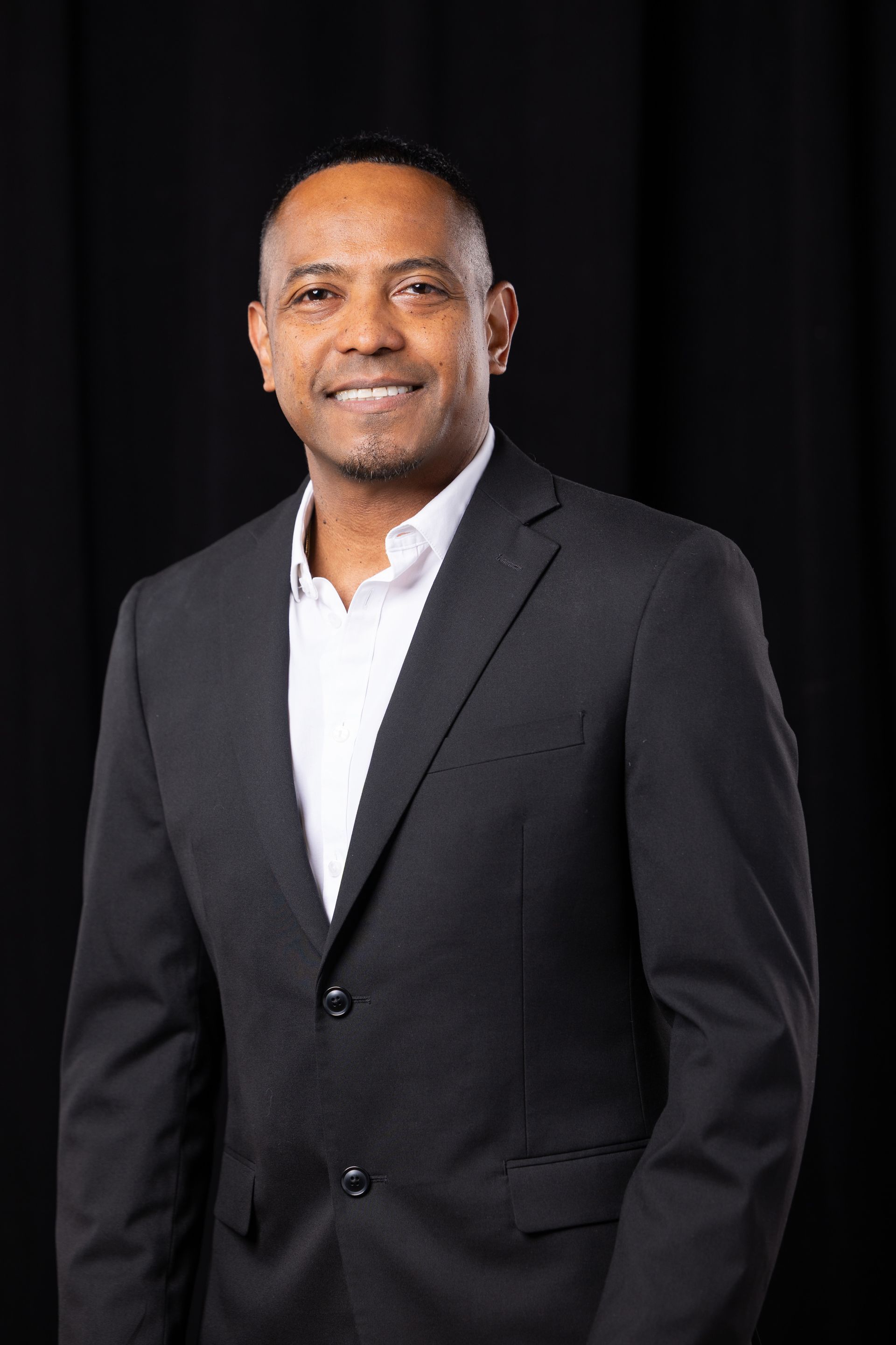 Man in a dark suit and white shirt standing against a black backdrop, smiling slightly
