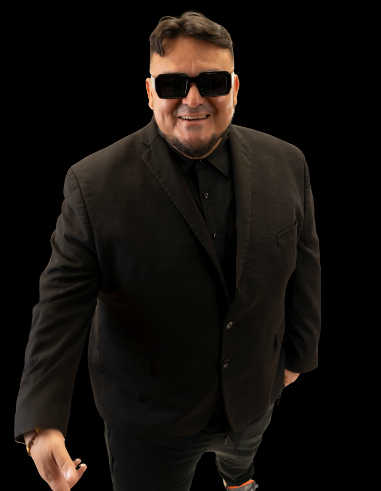 Smiling man in black suit and sunglasses standing against a black background.