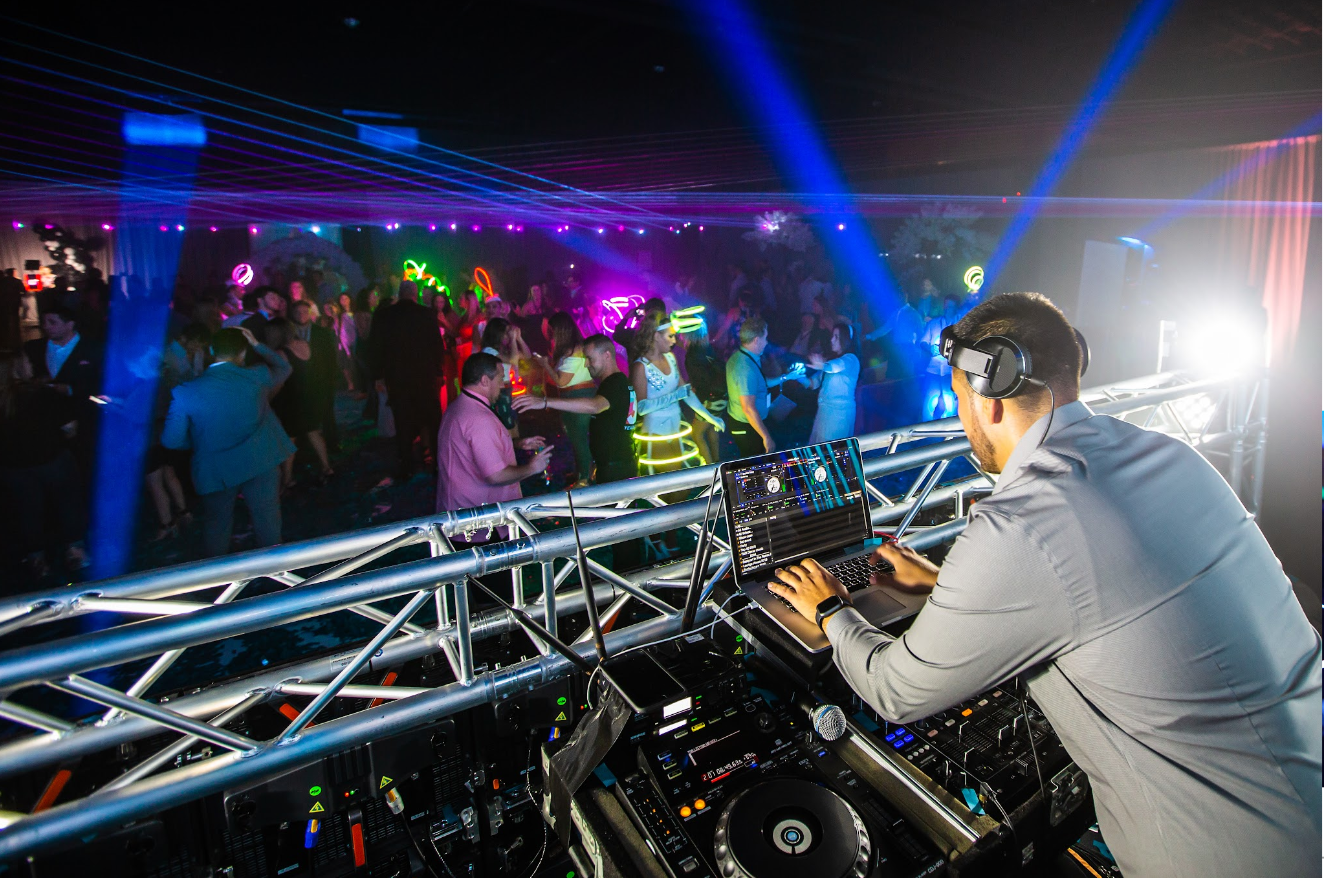 DJ performing at a nightclub, mixing music from behind a booth with colorful lights and crowd in the background