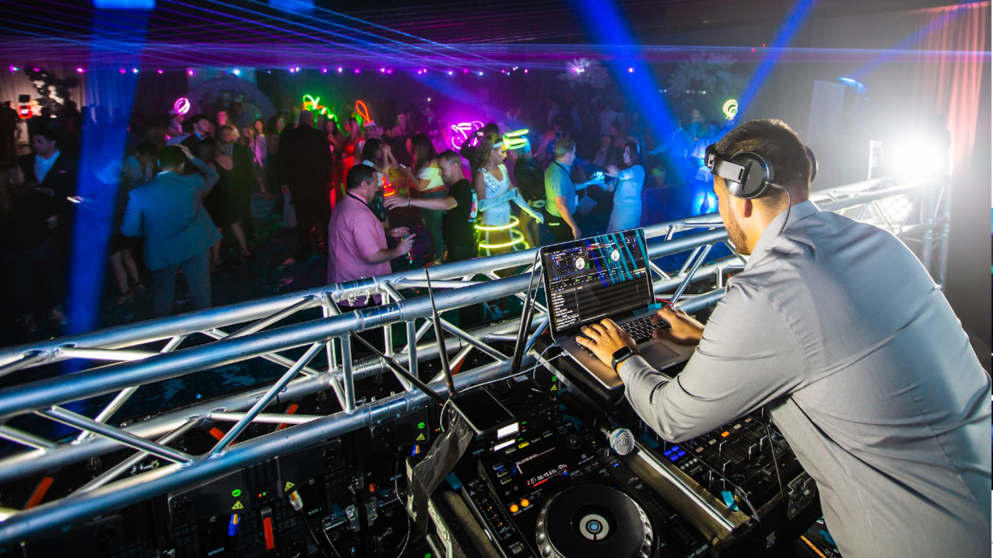 DJ performing on stage at a nightclub, mixing music under colorful lights and a crowd in the background