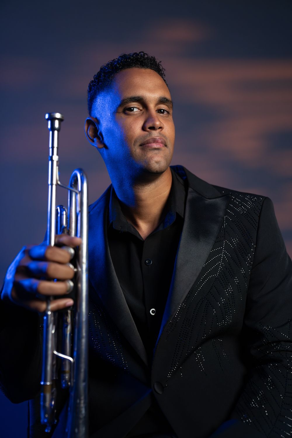 Smiling person in a black suit standing against a dark background, hands clasped in front.