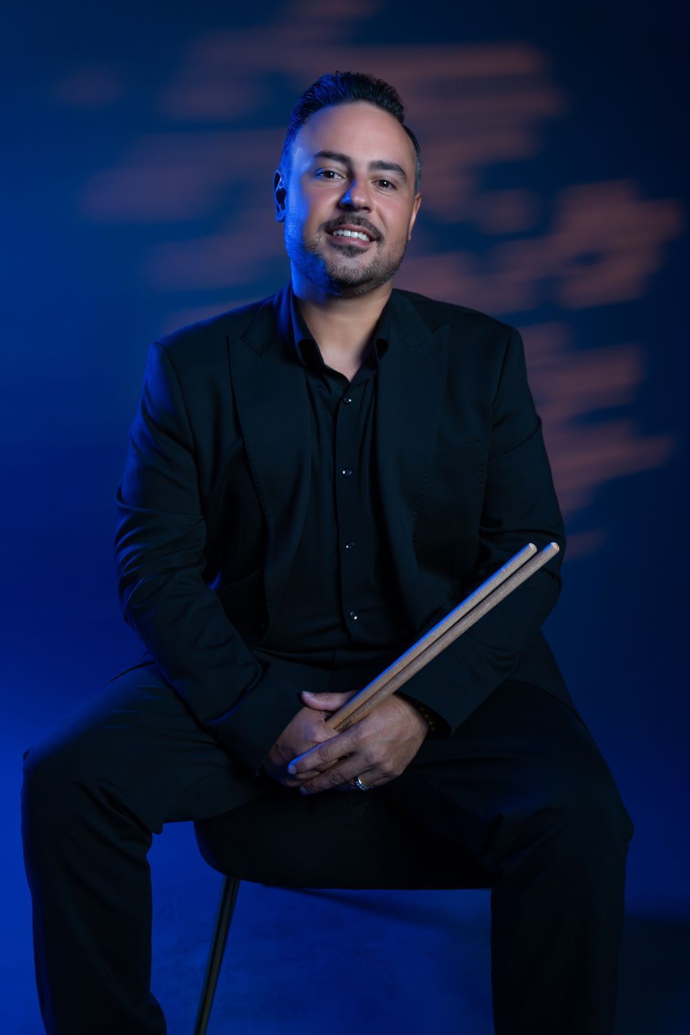 Smiling person in a black suit standing against a dark background, hands clasped in front.