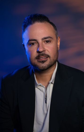 Man standing against a black background, wearing a black polo shirt and holding his hands together.