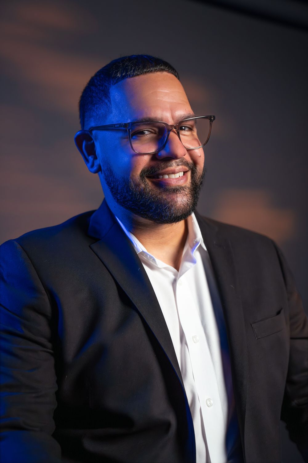Man in a black suit and white shirt standing against a black background, smiling with hands clasped.