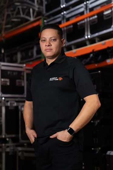 Man in a black polo shirt smiling against a dark background, hands clasped in front.