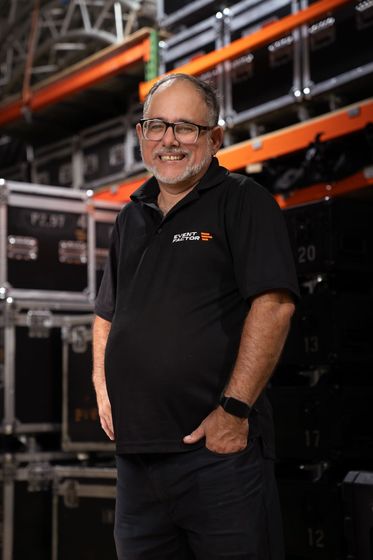 Man in black polo shirt and glasses standing against a black background, smiling with hands clasped.