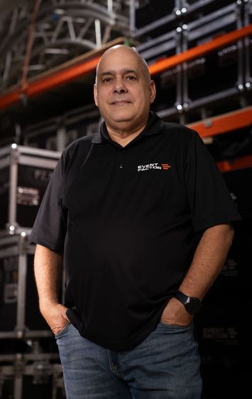 Man standing against a black background, wearing a black polo shirt and holding his hands together.