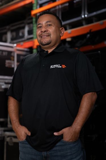 Man standing against a dark backdrop, wearing a black polo shirt with hands in pockets.