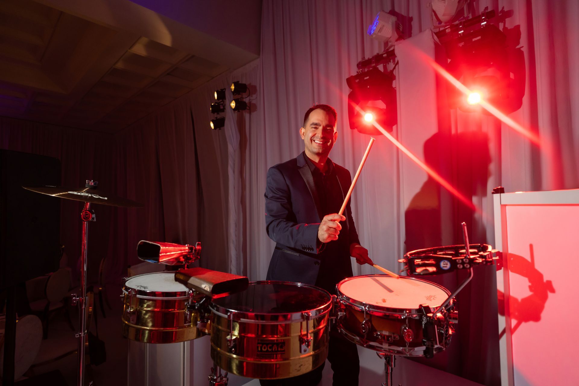 Musician playing timbales under red stage lights, holding drumsticks in a dim performance space