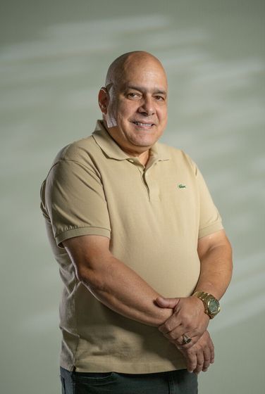 Man standing against a black background, wearing a black polo shirt and holding his hands together.
