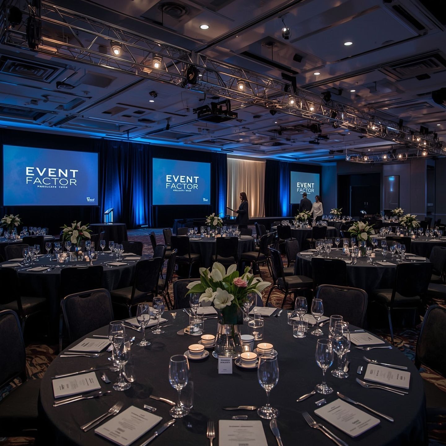 Blue-lit banquet hall set for an event, with round tables, glassware, and large presentation screens.