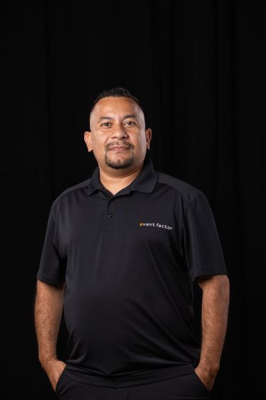 Man standing against a dark backdrop, wearing a black polo shirt with hands in pockets.