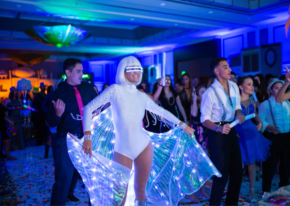 Person in a white bodysuit with glowing wings walking a runway under blue lights, with a crowd watching