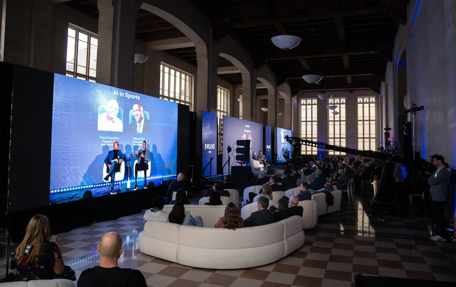 Conference audience watching a panel on a large blue-lit stage in a high-ceilinged hall