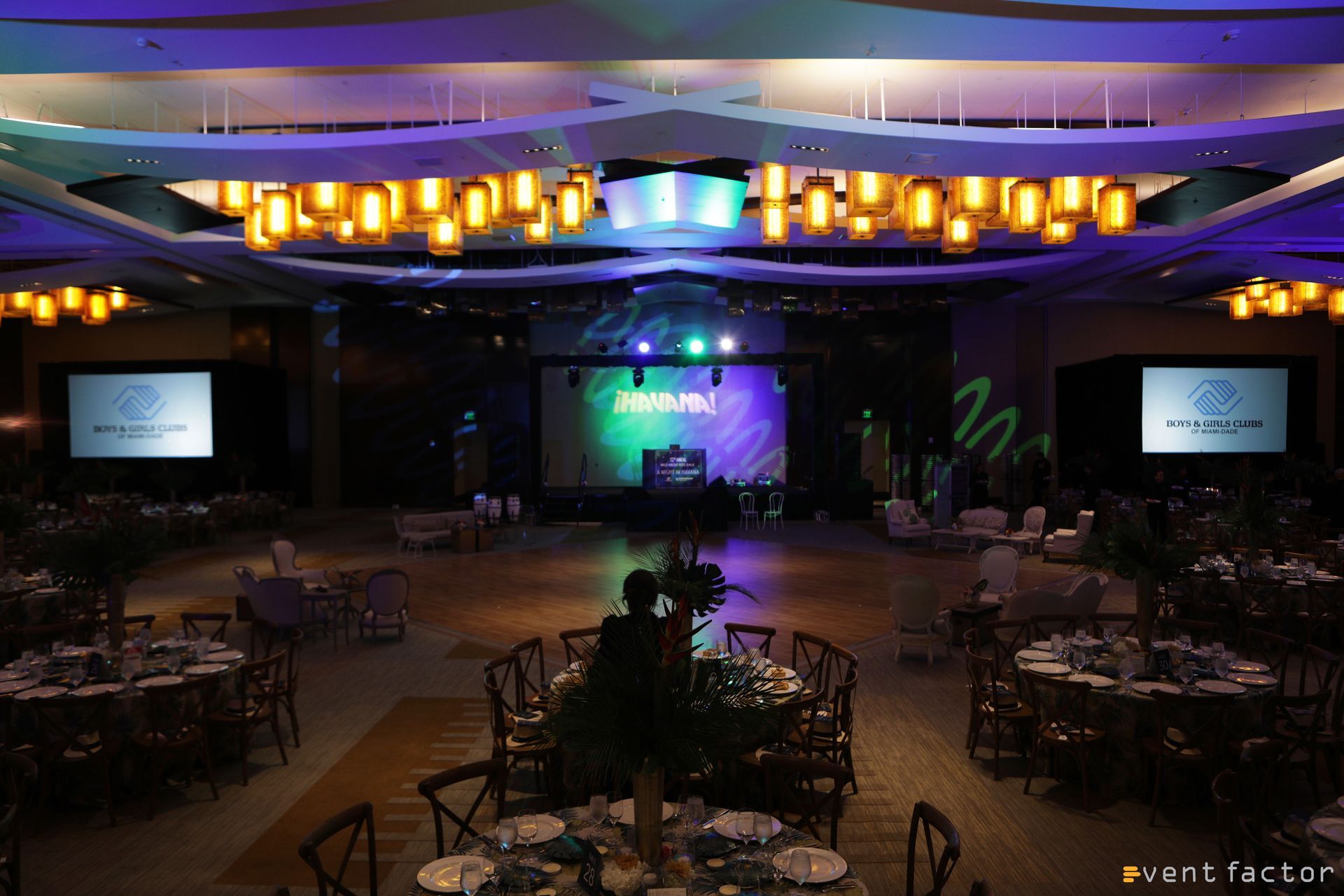 Dimly lit banquet hall with round tables, ceiling lights, and a stage with two projection screens.