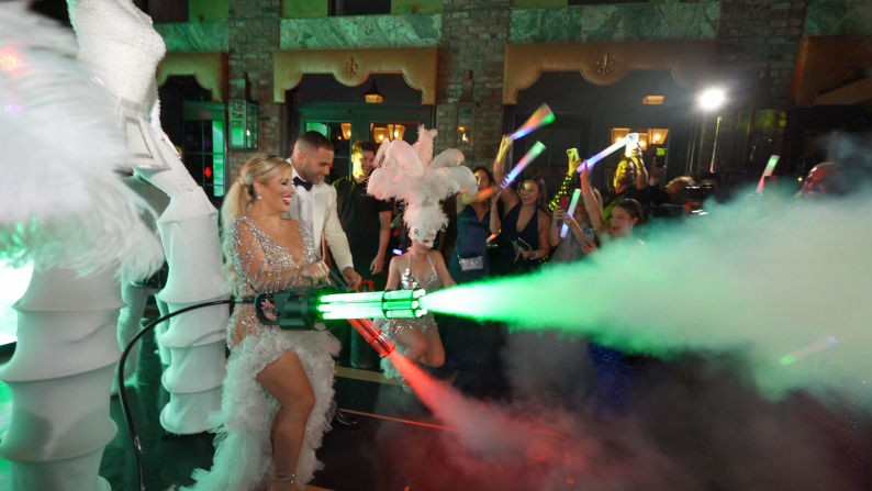 People in white costumes perform amid green fog and colorful lights at a lively indoor event