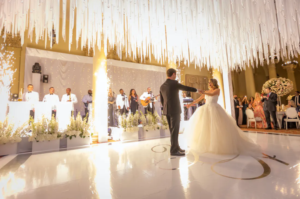 Couple dancing at a white-and-gold wedding reception with guests and decorative draped ceiling lights