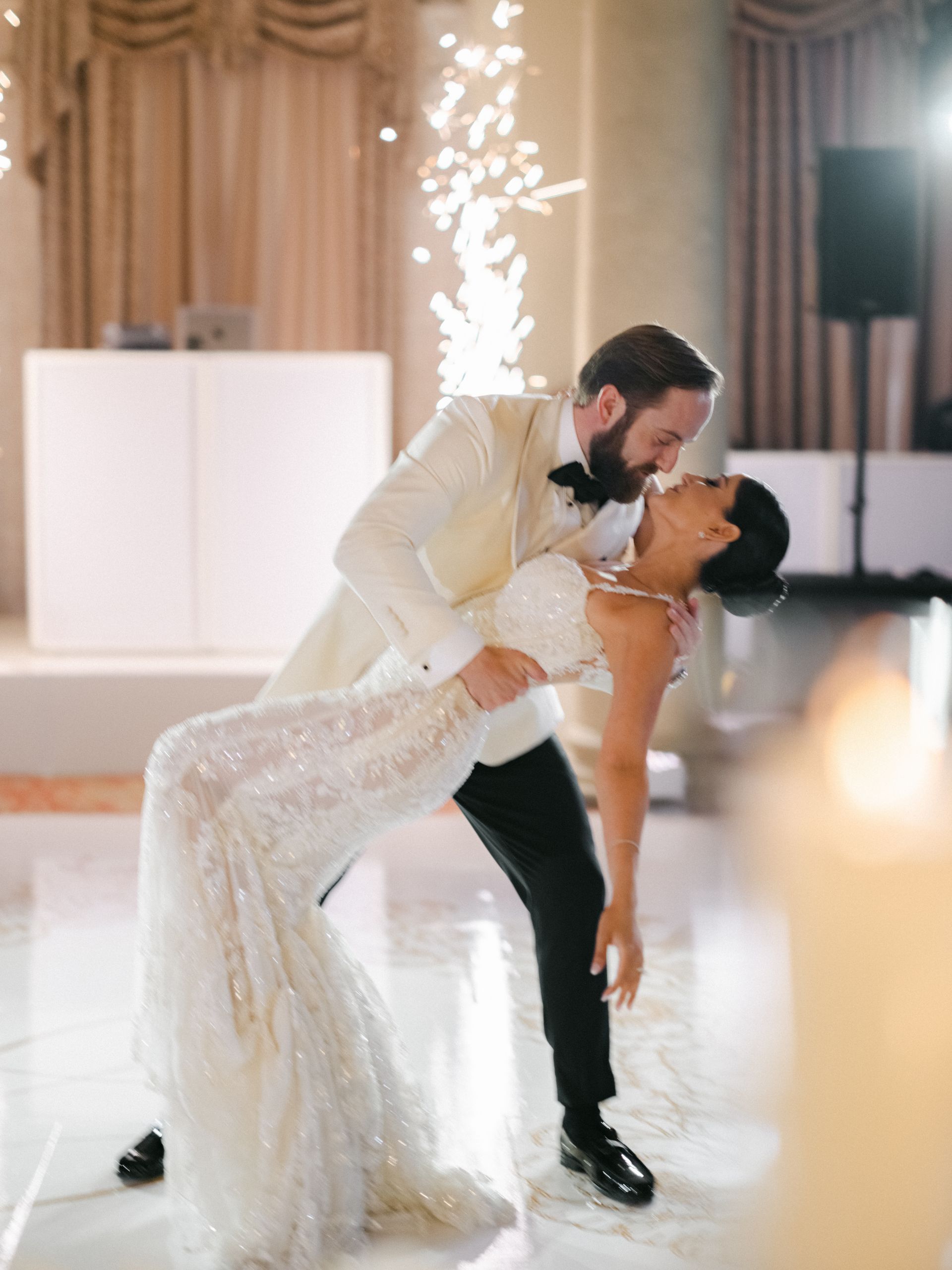 Bride and groom dancing in a white ballroom, groom dipping bride in a lace gown.