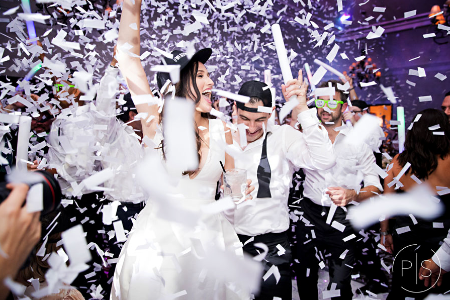 Confetti falls over a cheering couple in white outfits at a wedding celebration