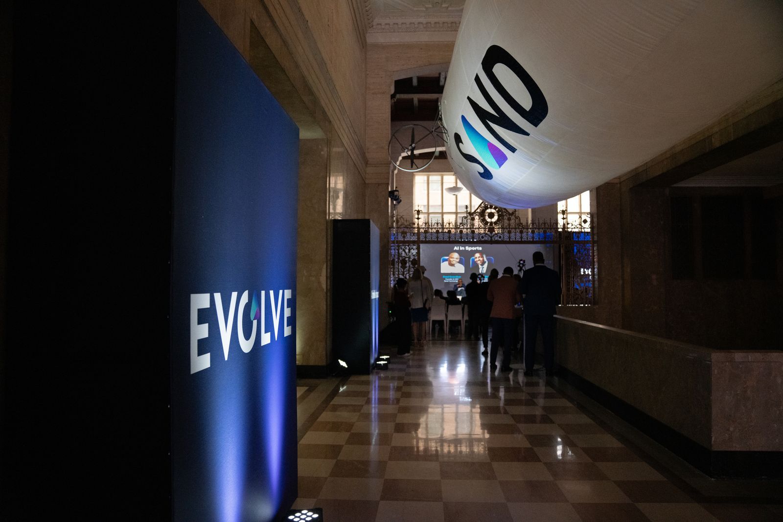 Dim hallway with an EVOLVE sign and a large hanging balloon, people gathered in the distance.