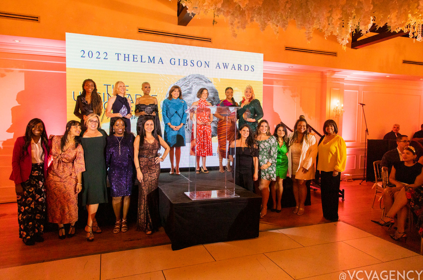 Women on a lit stage at an awards ceremony, posing with a banner backdrop and trophy pedestal