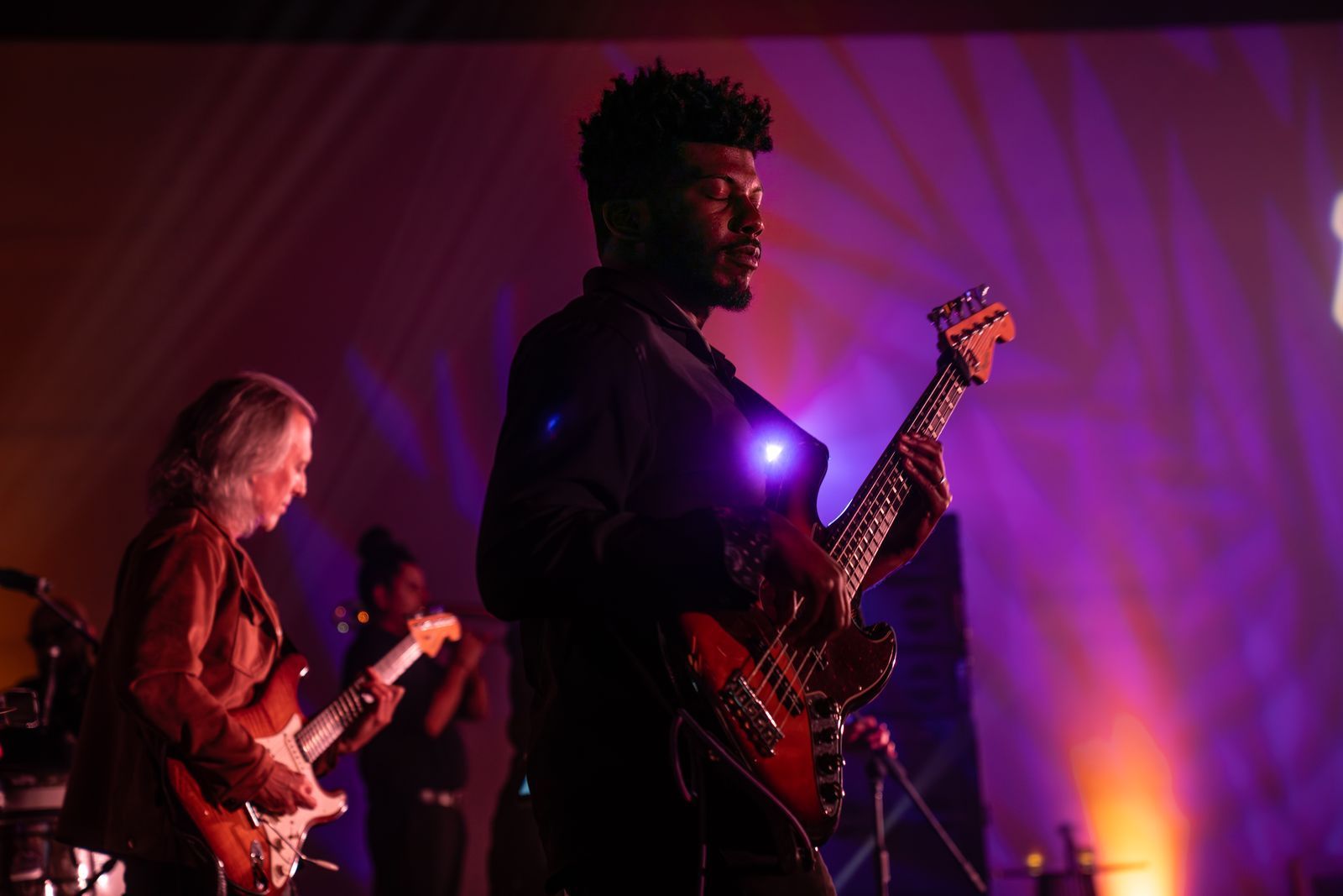 Musicians performing on stage under purple and orange lights, with guitars in the foreground
