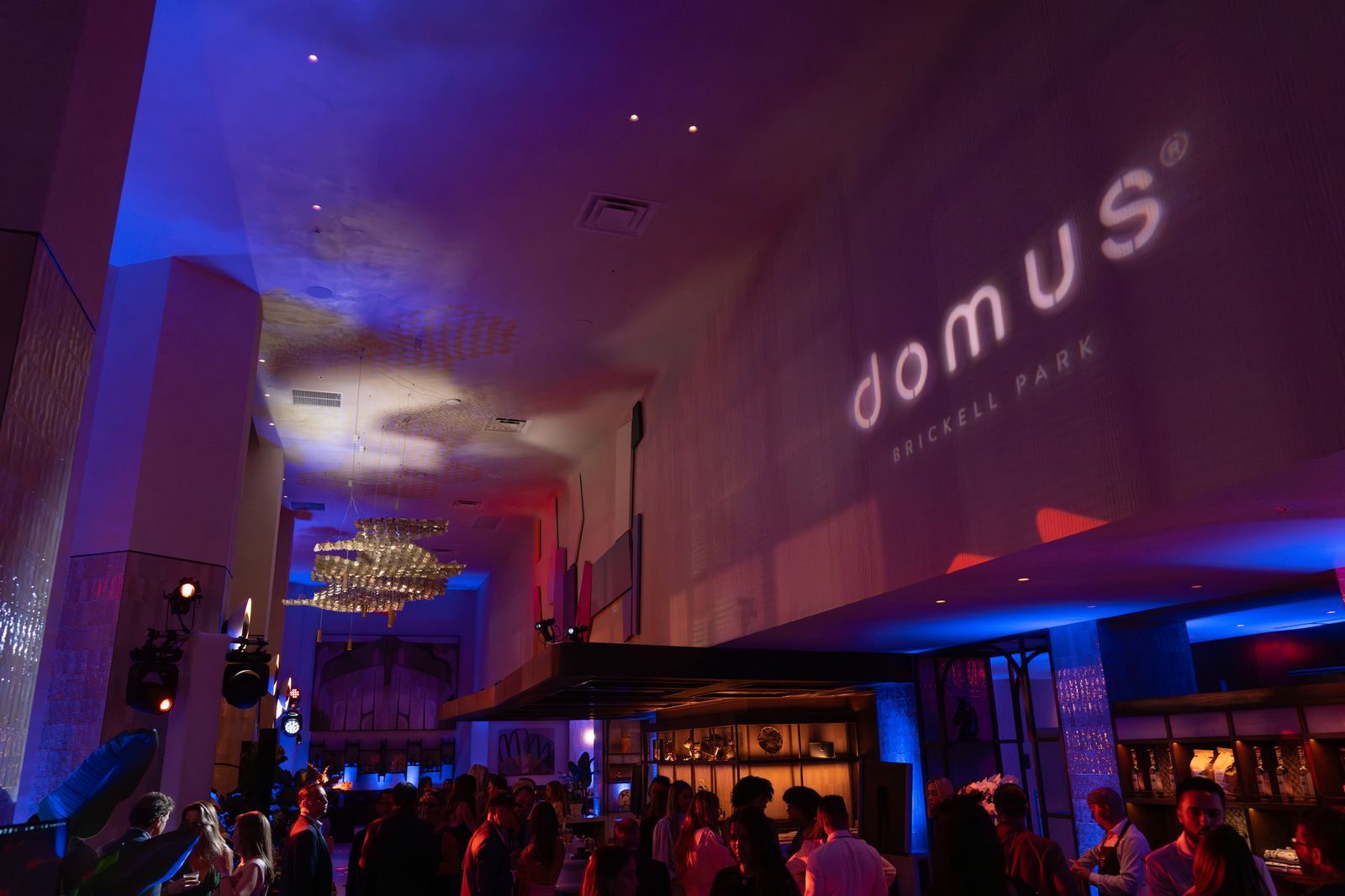 Crowded nightclub with purple-blue lighting and “domus” projected on the wall