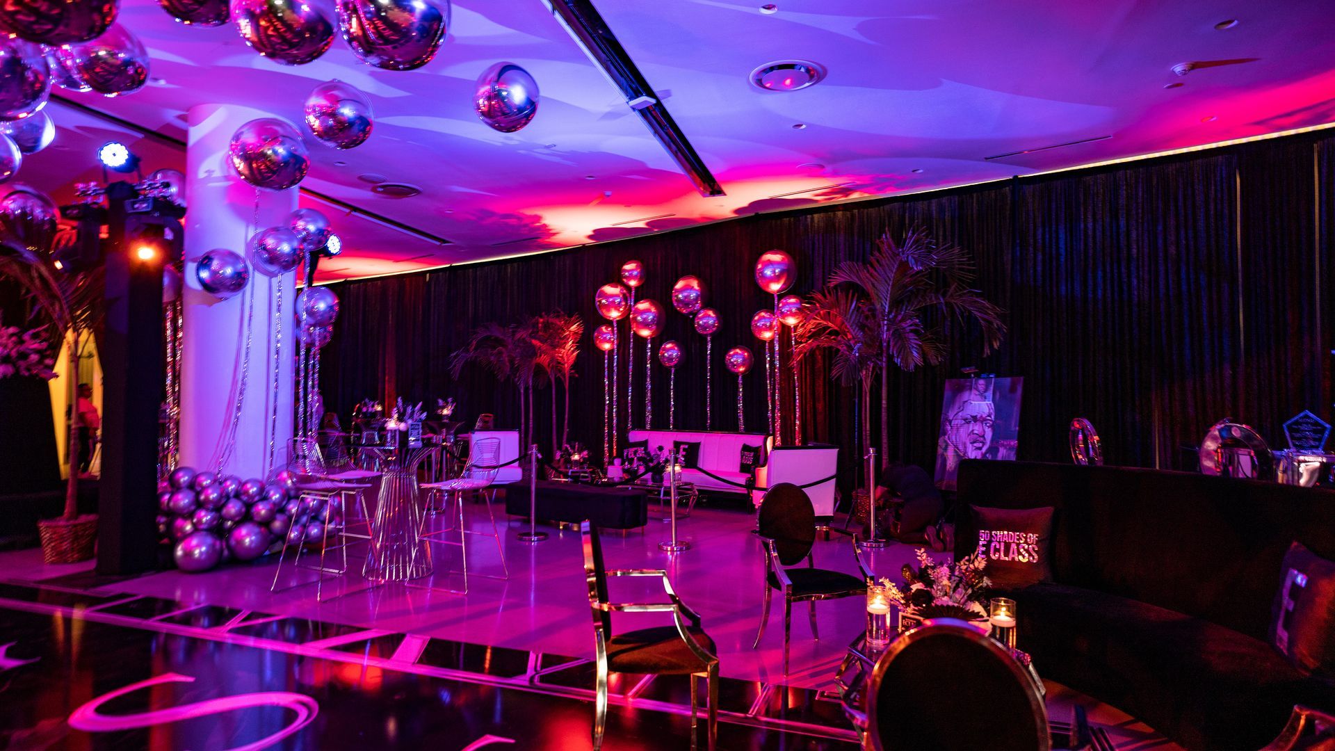Pink-lit event venue with tables, chairs, disco balls, and draped black curtains