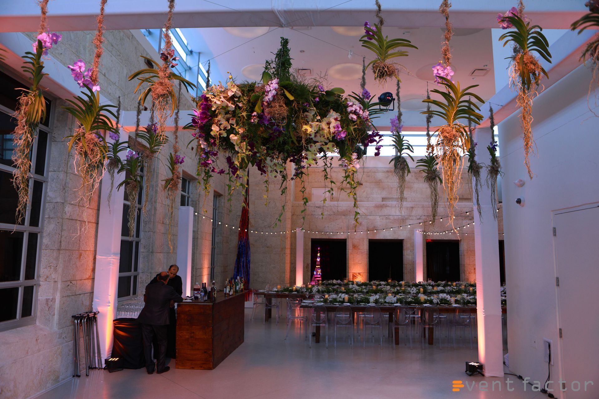Flower-filled indoor event space with hanging greenery, long tables, and soft pink lighting