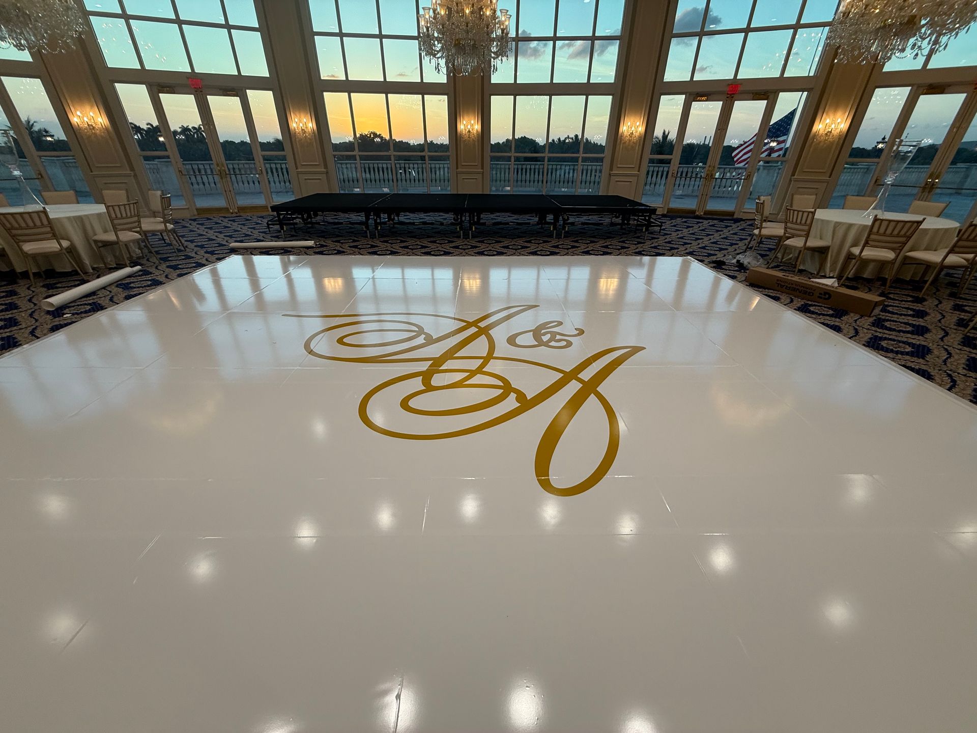 Elegant ballroom with a glossy white dance floor, gold monogram, and banquet chairs by large windows at sunset