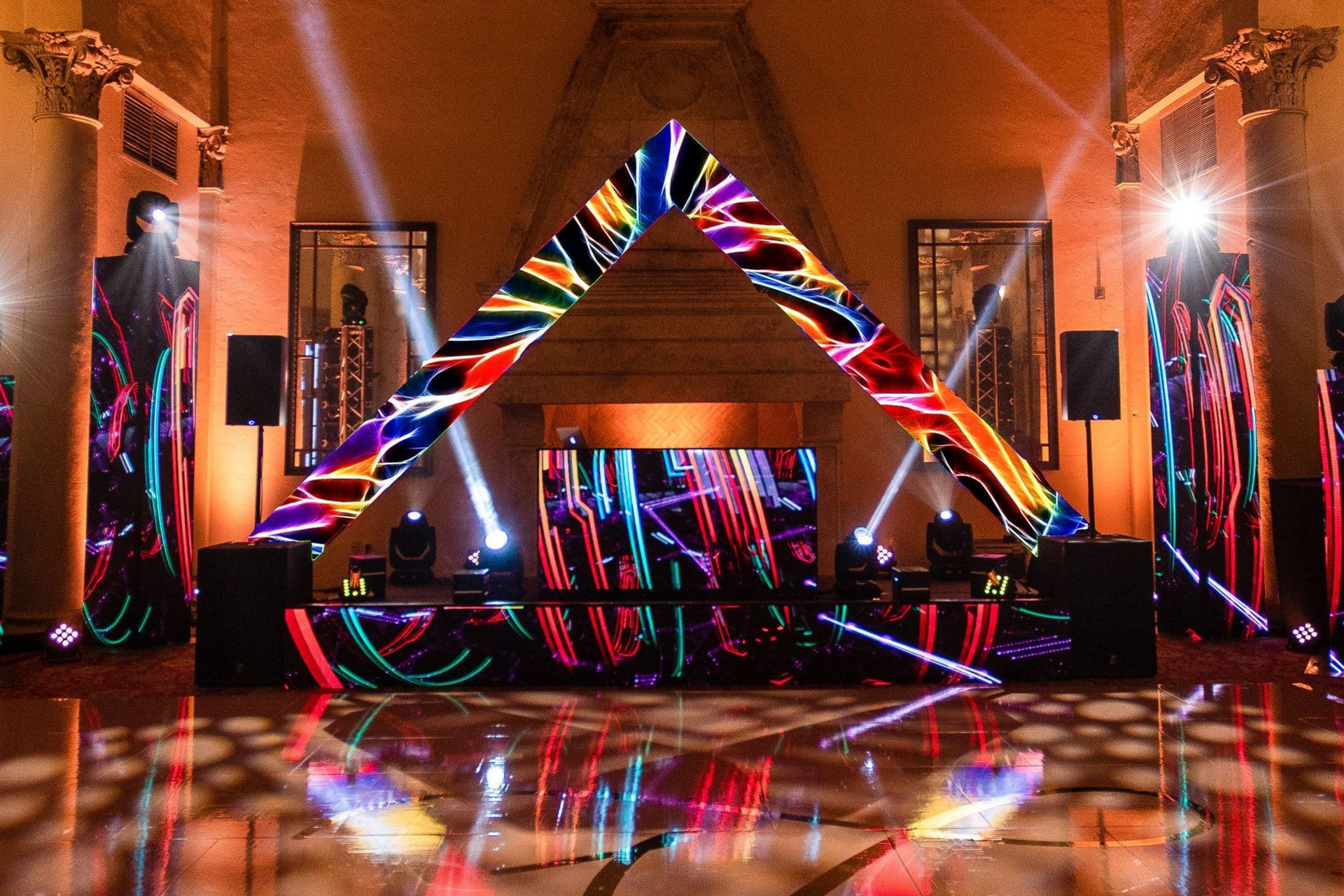 Colorful LED-lit DJ stage with geometric triangle backdrop in a warm indoor venue