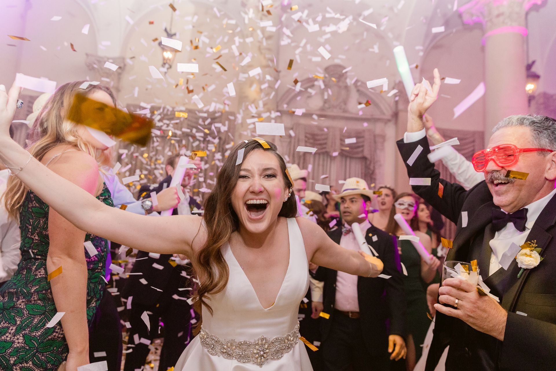 Wedding guests celebrating as confetti falls; a bride cheers in the center and a groom with pink glasses smiles on the right