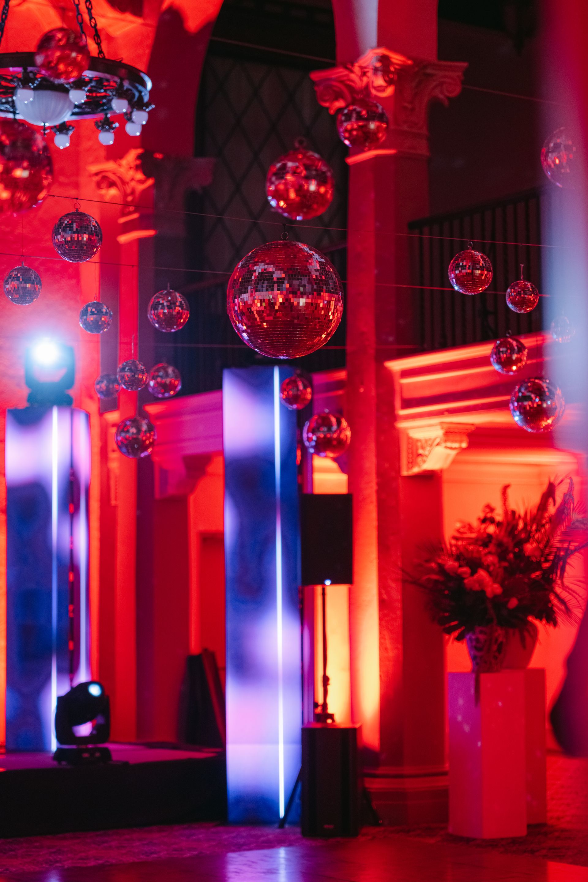 Red-lit indoor venue with hanging mirror balls, tall columns, and a potted plant by the wall.
