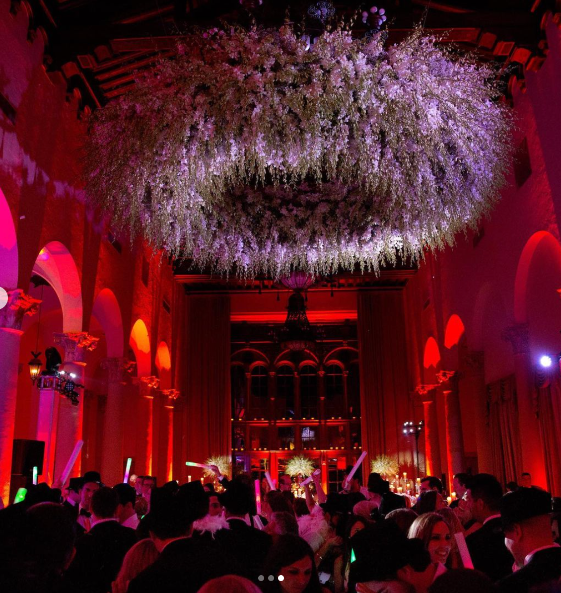 Crowded red-lit concert hall with a large hanging floral chandelier overhead