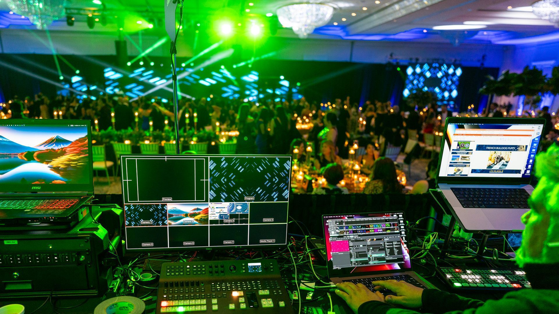 Green-lit control booth overlooking a crowded event, with multiple monitors and laptops in the foreground.