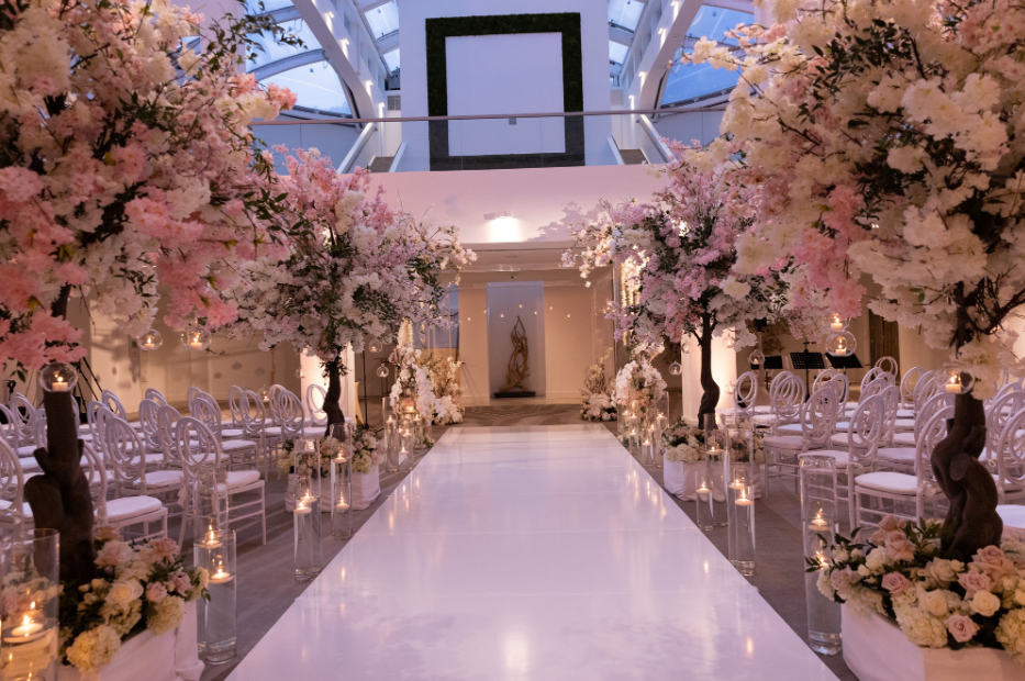 Elegant wedding aisle with pink floral arches, white chairs, and glossy white runway under soft lighting
