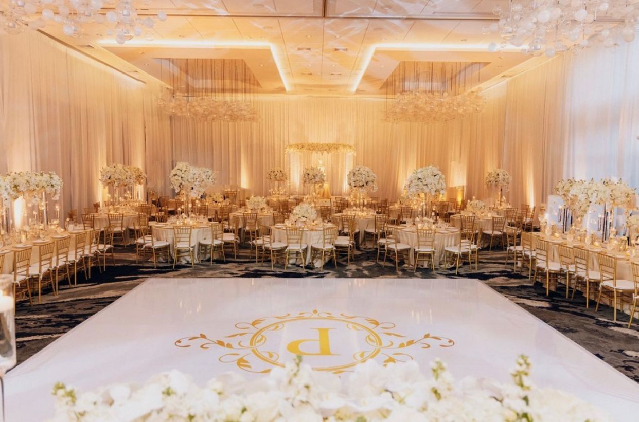 Elegant gold-and-white banquet hall with round tables, chandeliers, and a monogrammed dance floor