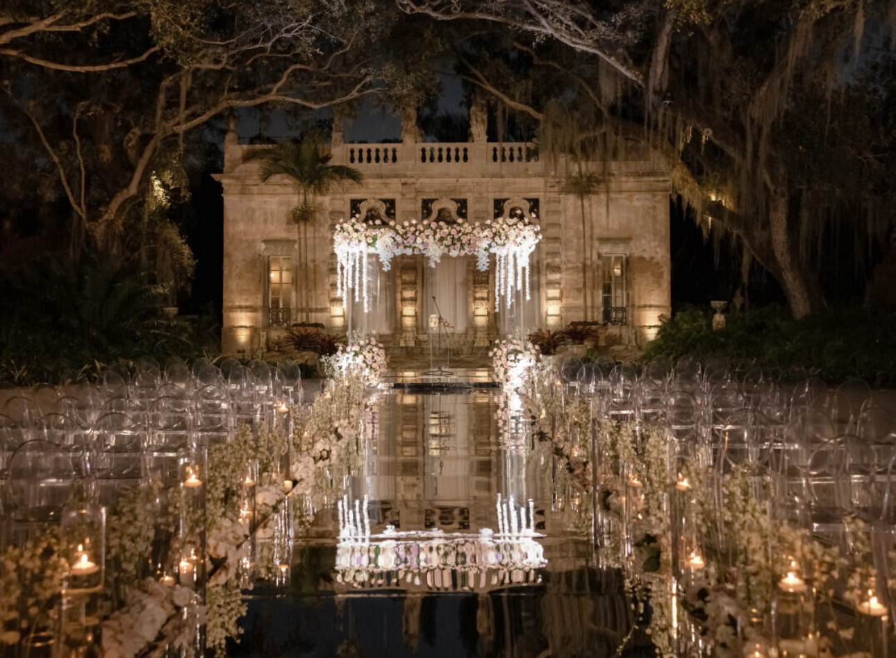 Nighttime view of a lit garden pavilion reflected in a still pool, surrounded by rows of candles and foliage