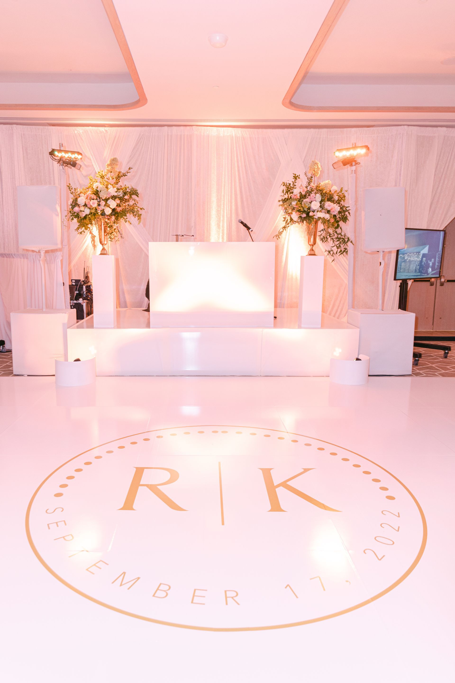 Elegant pink-lit wedding stage with white seating, floral arrangements, and monogrammed floor decal