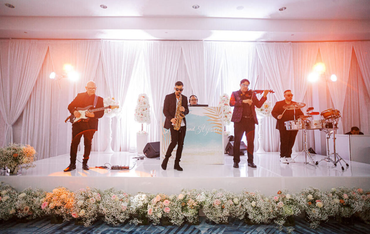 Live band performing on a white-draped stage with warm lights and floral arrangements in front