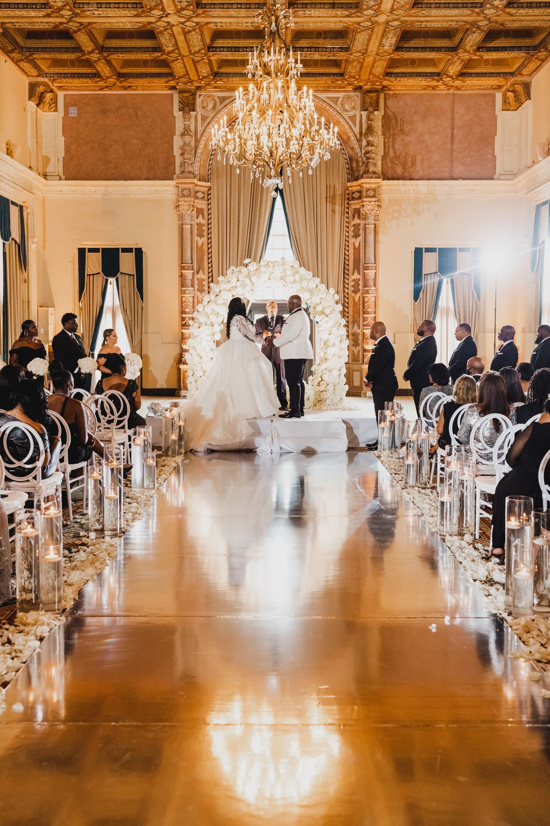 Elegant wedding ceremony in a grand gold ballroom, with bride and groom at the altar under a chandelier.
