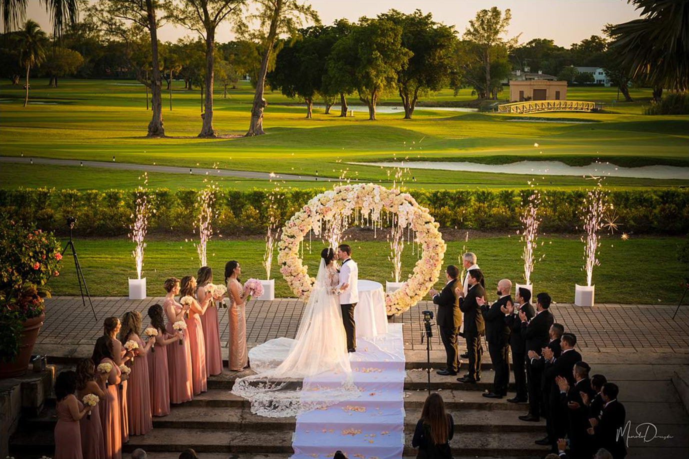 Wedding ceremony outdoors by a fountain, with bride and groom beneath a floral arch and wedding party lined up on both sides