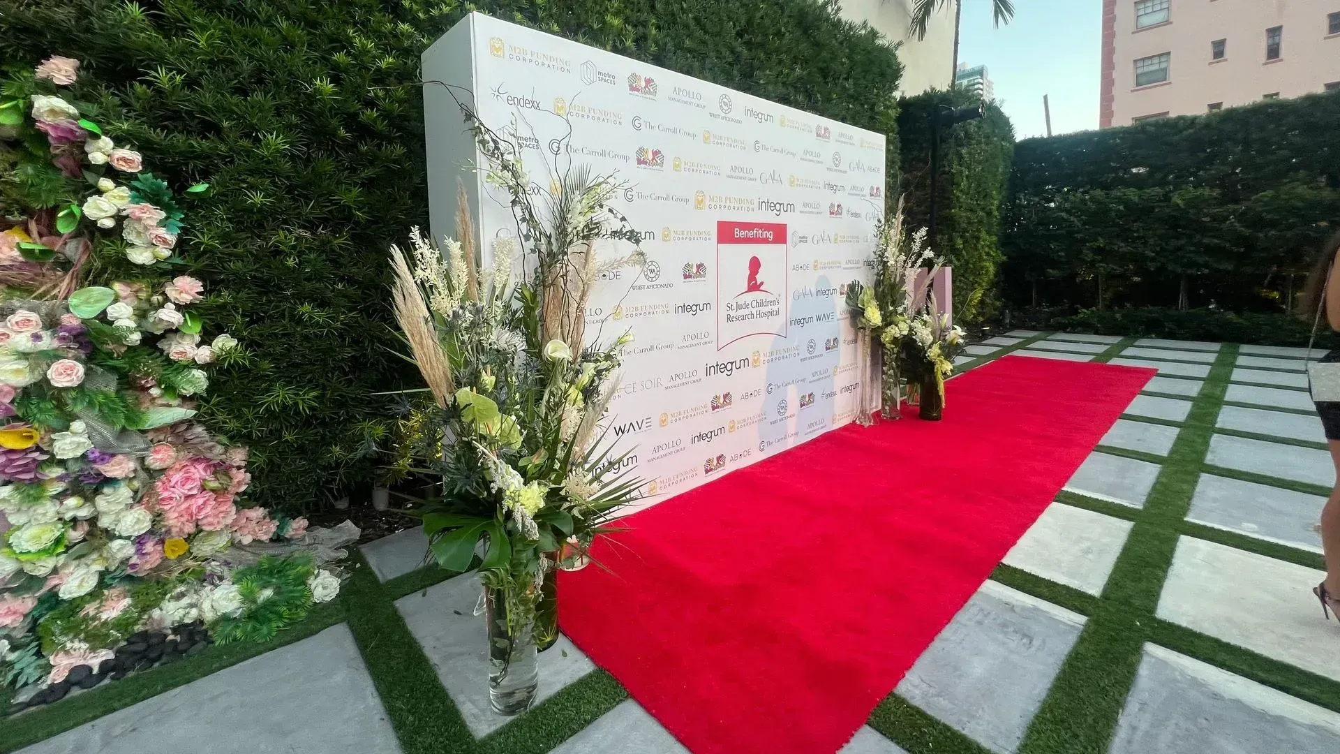 Red carpet beside a white event backdrop and floral display in an outdoor garden setting