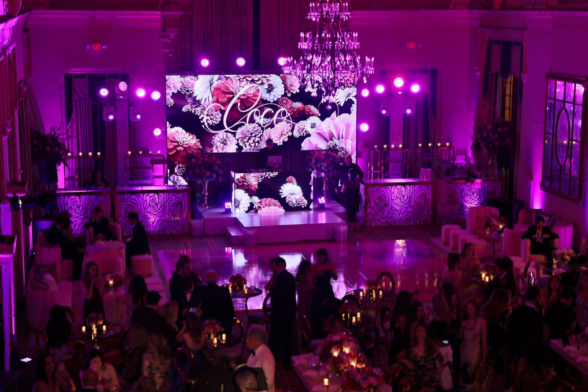Purple-lit banquet hall with guests seated at tables facing a large stage and screen showing floral graphics